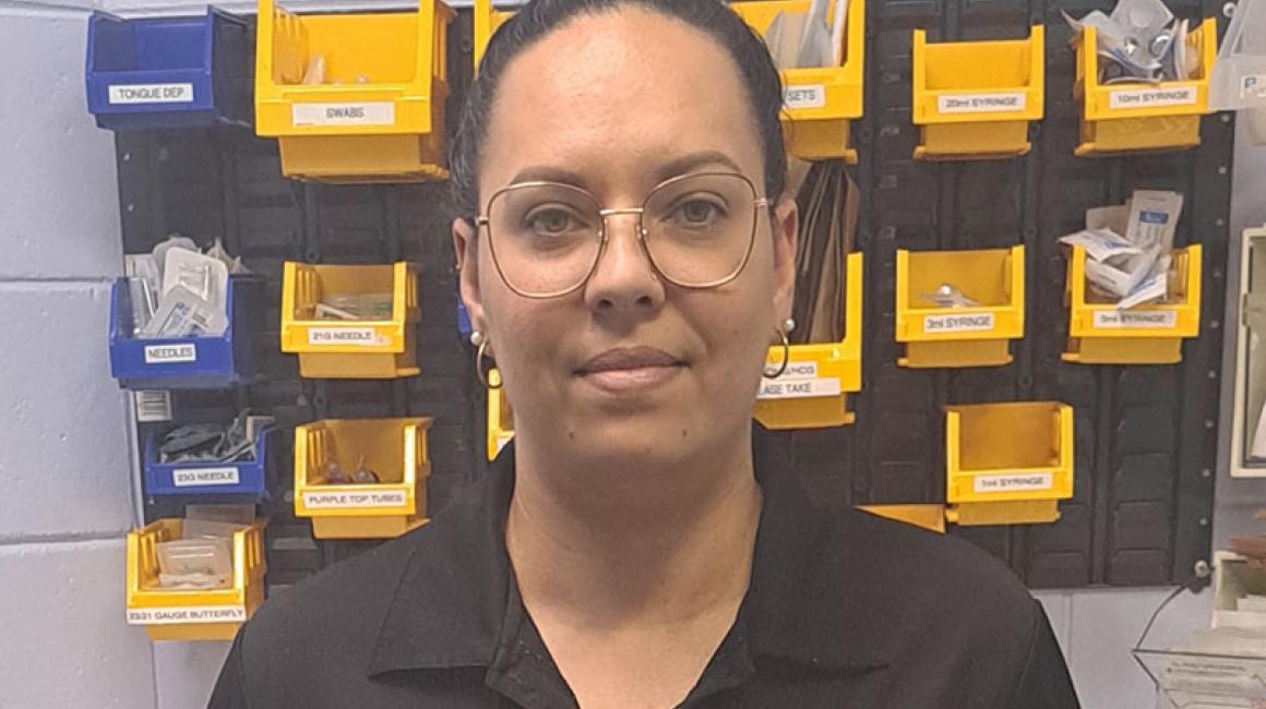 Cameilla is wearing square wire frame glasses and looking at the camera. She has a black Danila Dilba polo, dark hair and is standing in front of clinic medical supplies. 