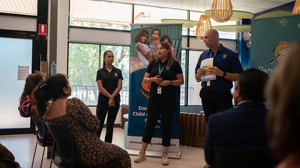 Casey is wearing a black Danila Dilba polo and standing in between an AHP and Danila Dilba CEO Rob McPhee. She is giving a speech to an audience at Darrandirra Child and Family Cente.